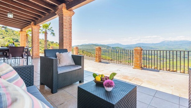 Eine Terrasse im Freien mit Korbmöbeln, einem Tisch mit einem Blumentopf und Blick auf ferne Hügel.