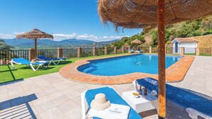 Ein Pool mit blauen Liegestühlen und einem Reetdachschirm, umgeben von einer Steindeck und Blick auf Berge.