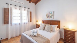 Ein ordentlich eingerichtetes Schlafzimmer mit einem Bett, zwei Nachttischen, einem Kopfteil, einem Gemälde und einem Fenster mit Vorhängen.