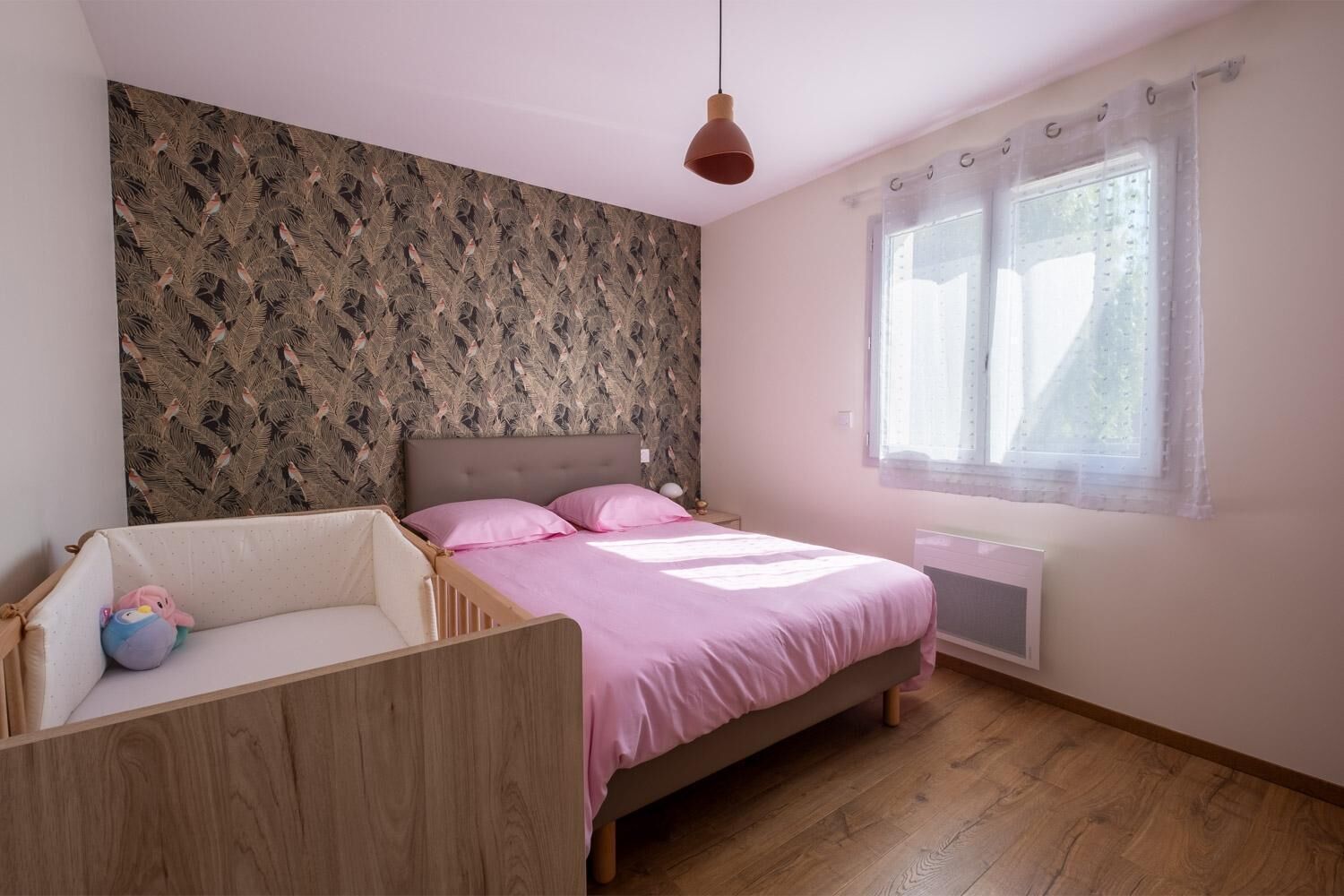 Ein Schlafzimmer mit einem Bett, einem Babybett, einem Fenster mit Vorhängen und einer Wand mit einem Blattmuster.