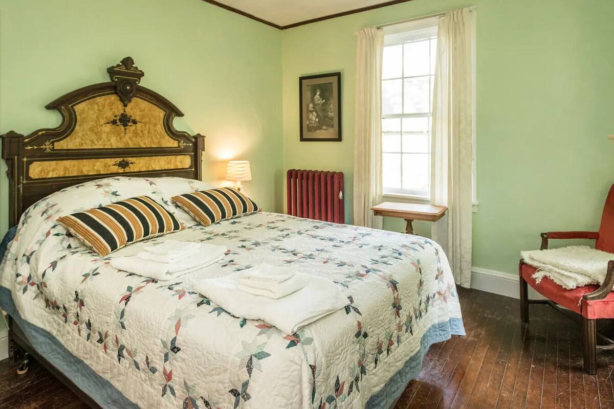 A bedroom with a large bed, a wooden headboard, a red chair, a window with curtains, and a radiator.