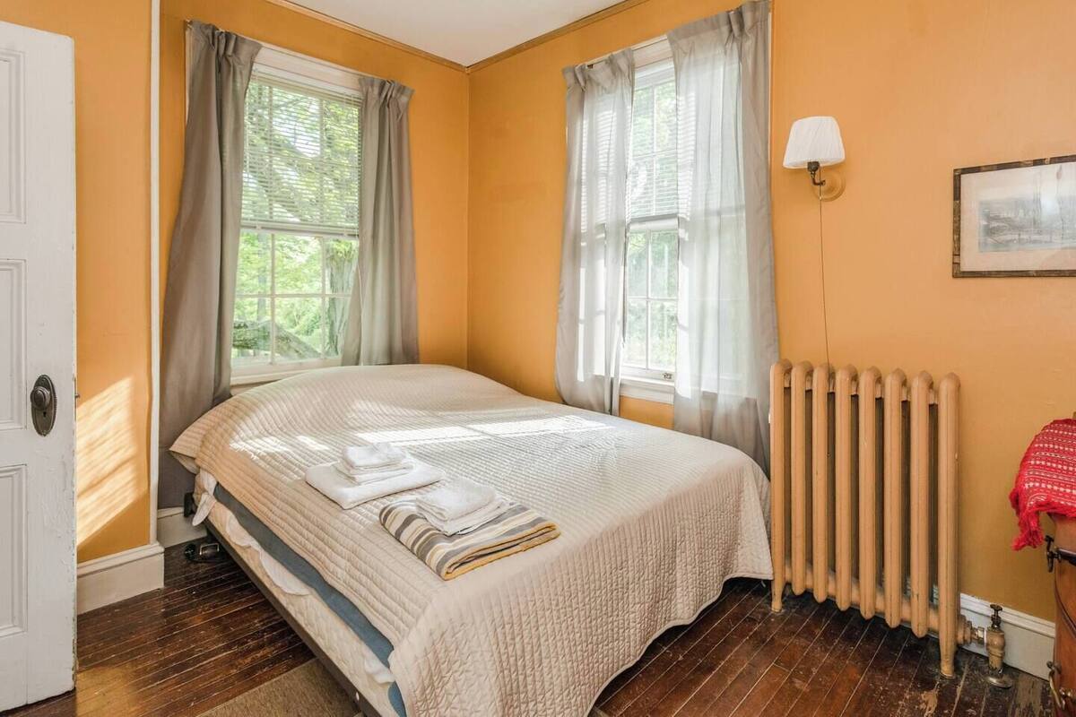 A bedroom with a bed, two windows with curtains, a radiator, and a wall-mounted lamp.