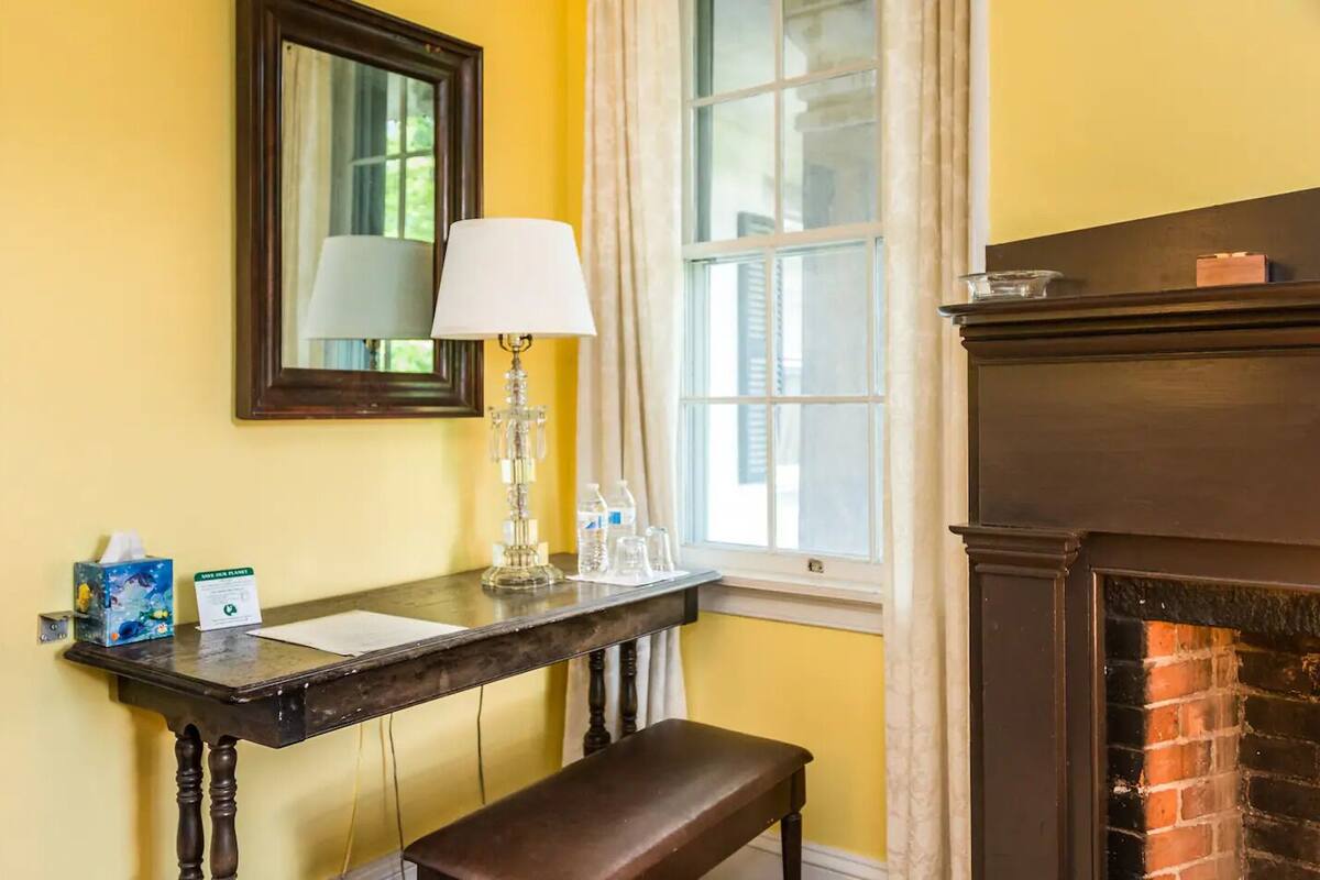 A room with a wooden bench, a table with a lamp, a mirror, and a fireplace.