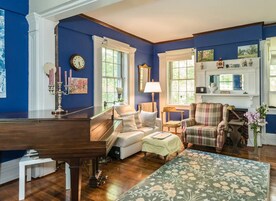 Uma sala de estar com um piano, um sofá, uma poltrona xadrez e uma lareira.