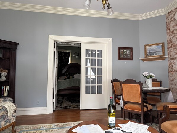 A room with a wooden floor, a dining table with chairs, a bookshelf, and a door leading to another room. - The Gallery (Hermann)
