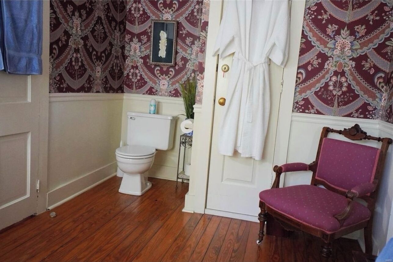 Een badkamer met een toilet, een witte deur, een houten stoel en behang met een patroon.