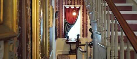 A grand hallway with ornate wall decorations, a wooden staircase with white balusters, and a red curtain at the end.