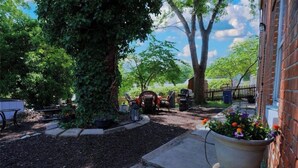 A garden area with a brick wall, a large tree, and a potted plant with flowers. - The Gallery (Hermann)