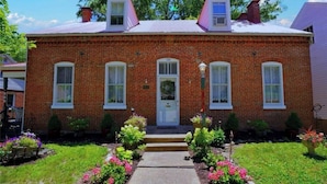 A two-story brick house with a front porch, a front door, and a small garden with flowers and shrubs. - The Gallery (Hermann)