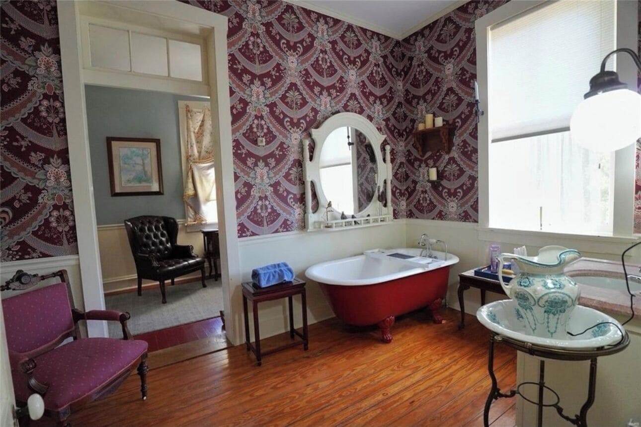 A bathroom with a red clawfoot bathtub, a white sink, and a mirror above it. There is a window with a white lamp beside it.