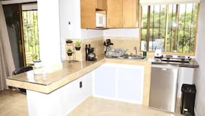 A compact kitchen with a microwave, stove, and sink. There is a small refrigerator and a trash bin. The kitchen has a window with a view of greenery outside.
