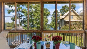 Une terrasse en bois avec une table et des chaises, de grandes fenêtres donnant sur des arbres et une maison, et des plantes en pot sur la table.