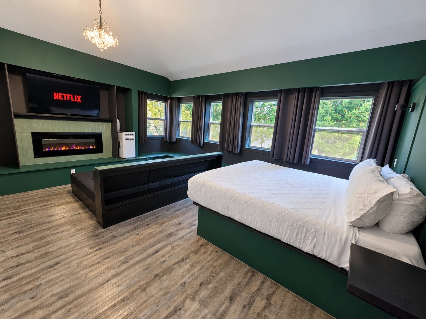 A modern bedroom with a large bed, a flat-screen TV mounted above a fireplace, and a view of greenery through the windows.