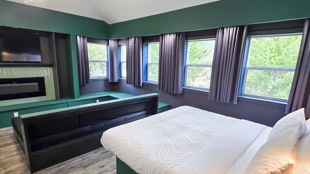 A bedroom with a bed, a built-in bench, a TV, and large windows. - Copper Rose Retreat B&B (Invermere)