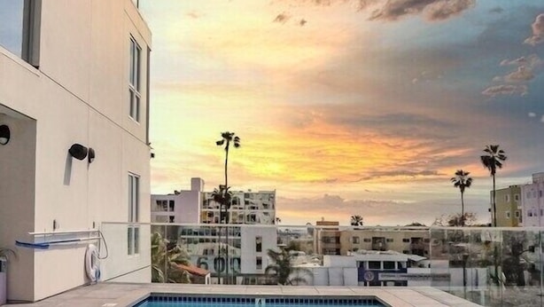 A rectangular swimming pool with clear blue water, surrounded by a tiled deck. In the background, there are modern buildings with balconies and a sunset sky with scattered clouds. - Hollywood sign view/ 5 mins from universal studio (West Hollywood)