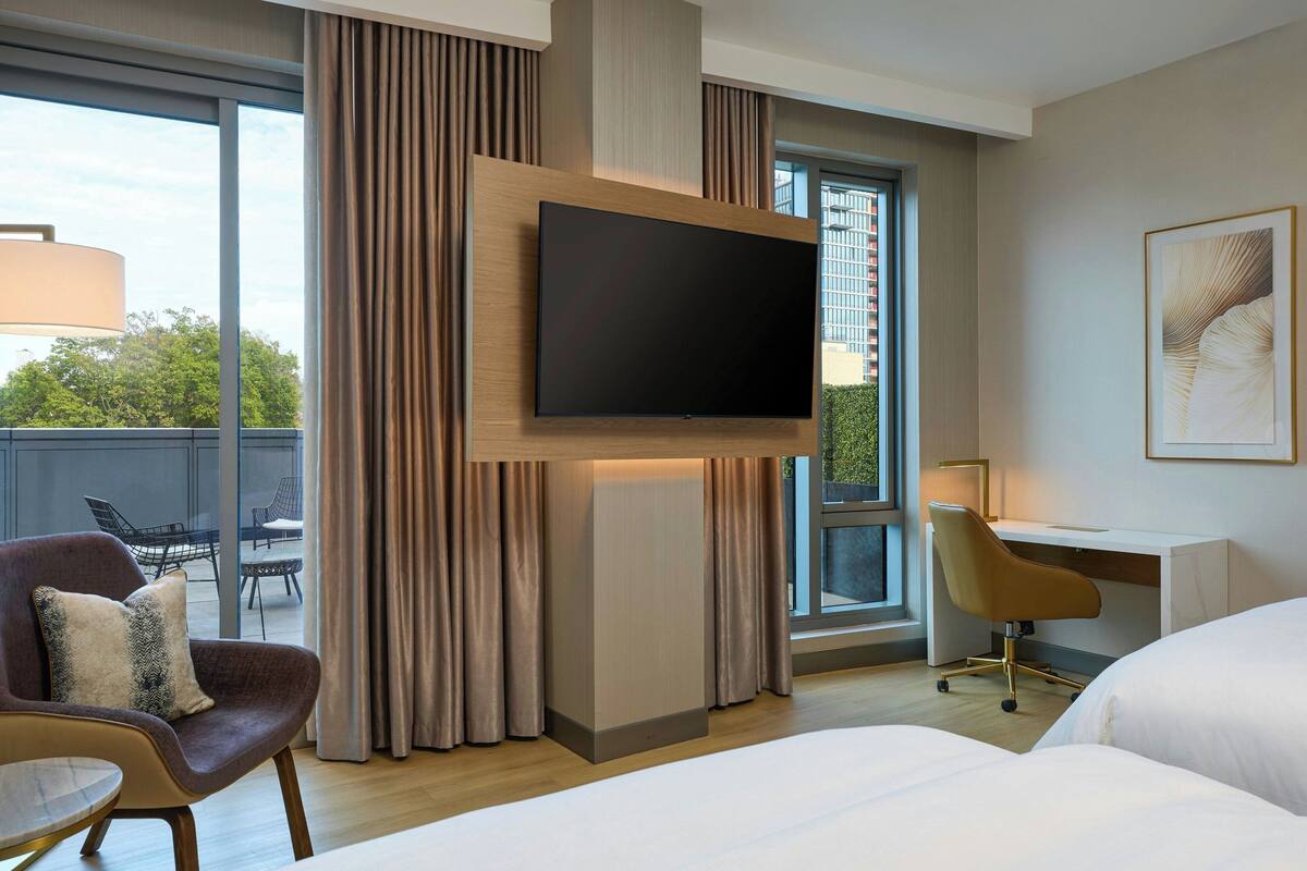 A modern hotel room with a large flat-screen TV mounted on the wall, a comfortable seating area with a chair and a small table, a desk with a chair, and a large bed.