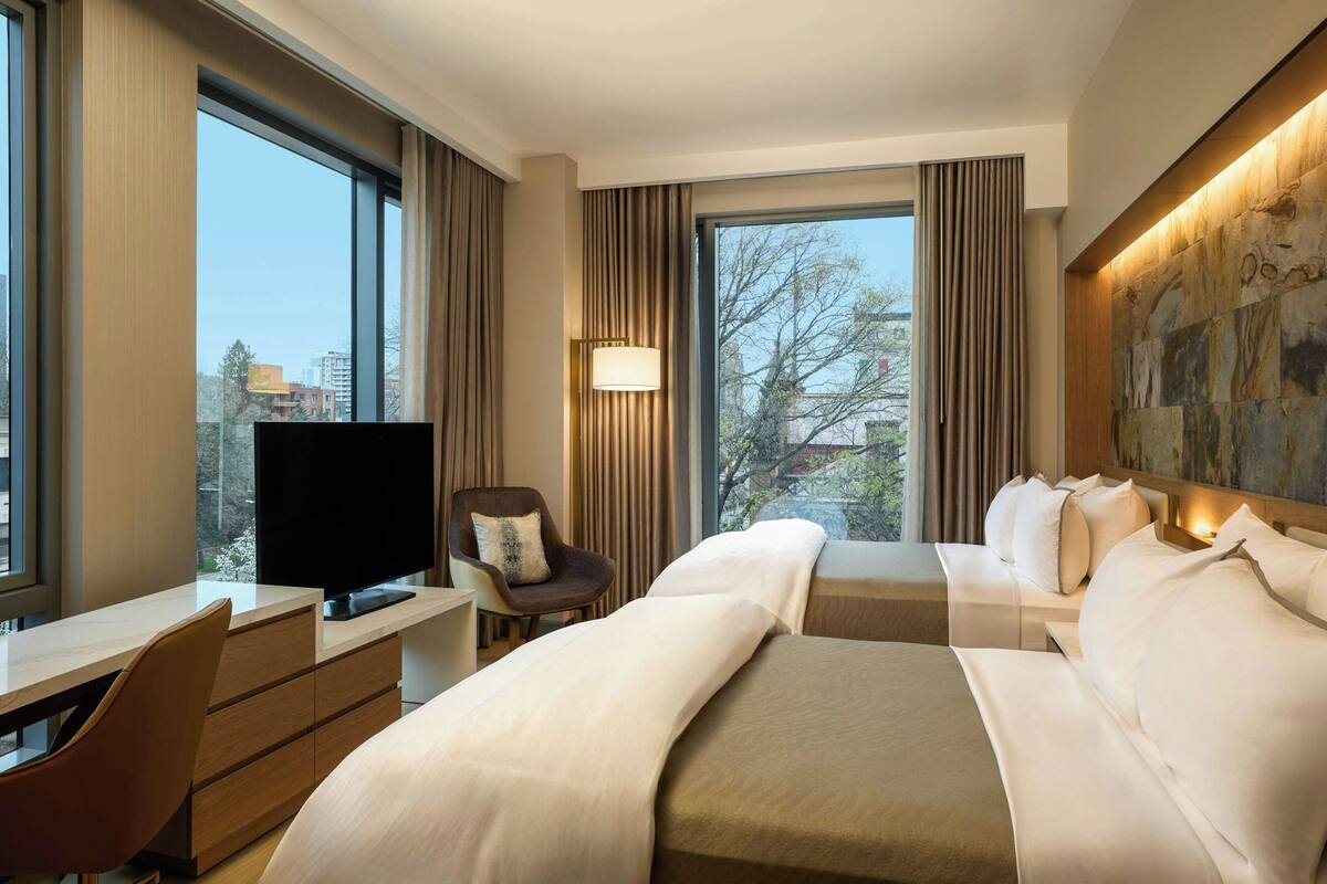 A hotel room with a large bed, a desk with a TV, a chair, and a view of the city.