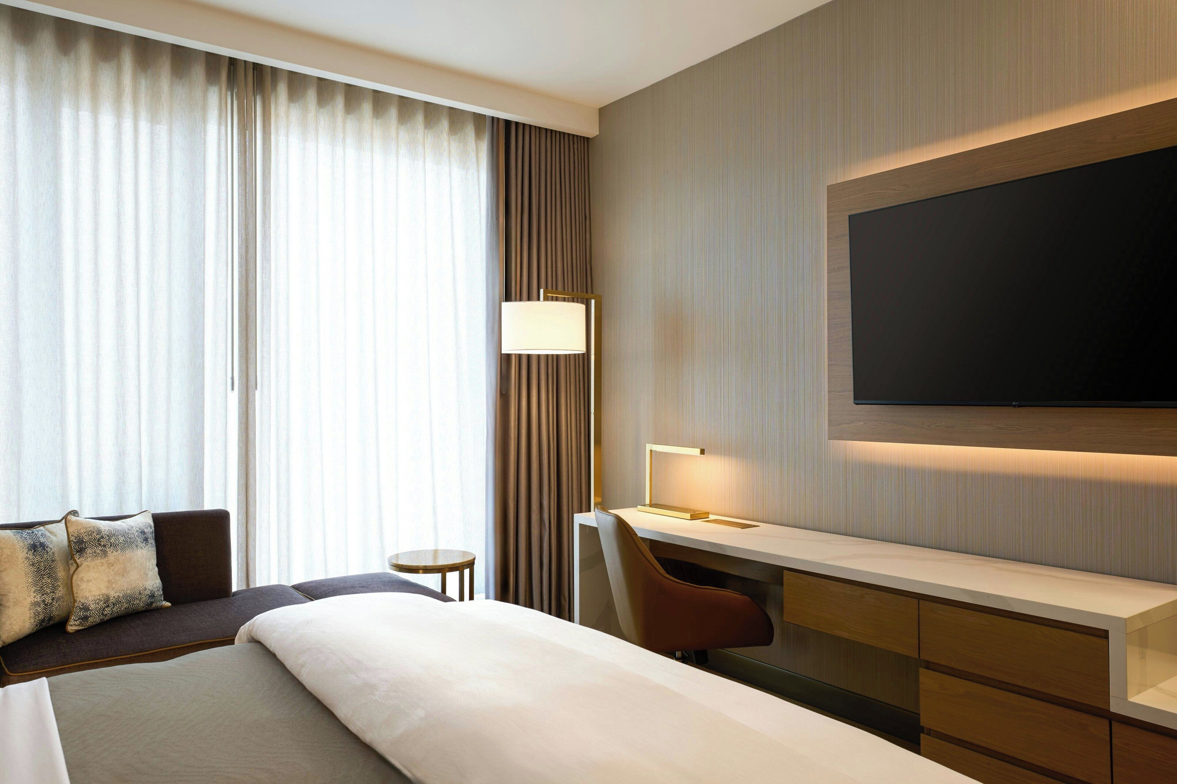 A hotel room with a large bed, a desk with a chair, a TV mounted on the wall, and a window with curtains.
