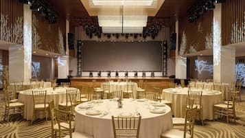 Uma sala de conferências com várias mesas redondas, cada uma disposta com cadeiras e utensílios de mesa, e uma tela grande na frente.