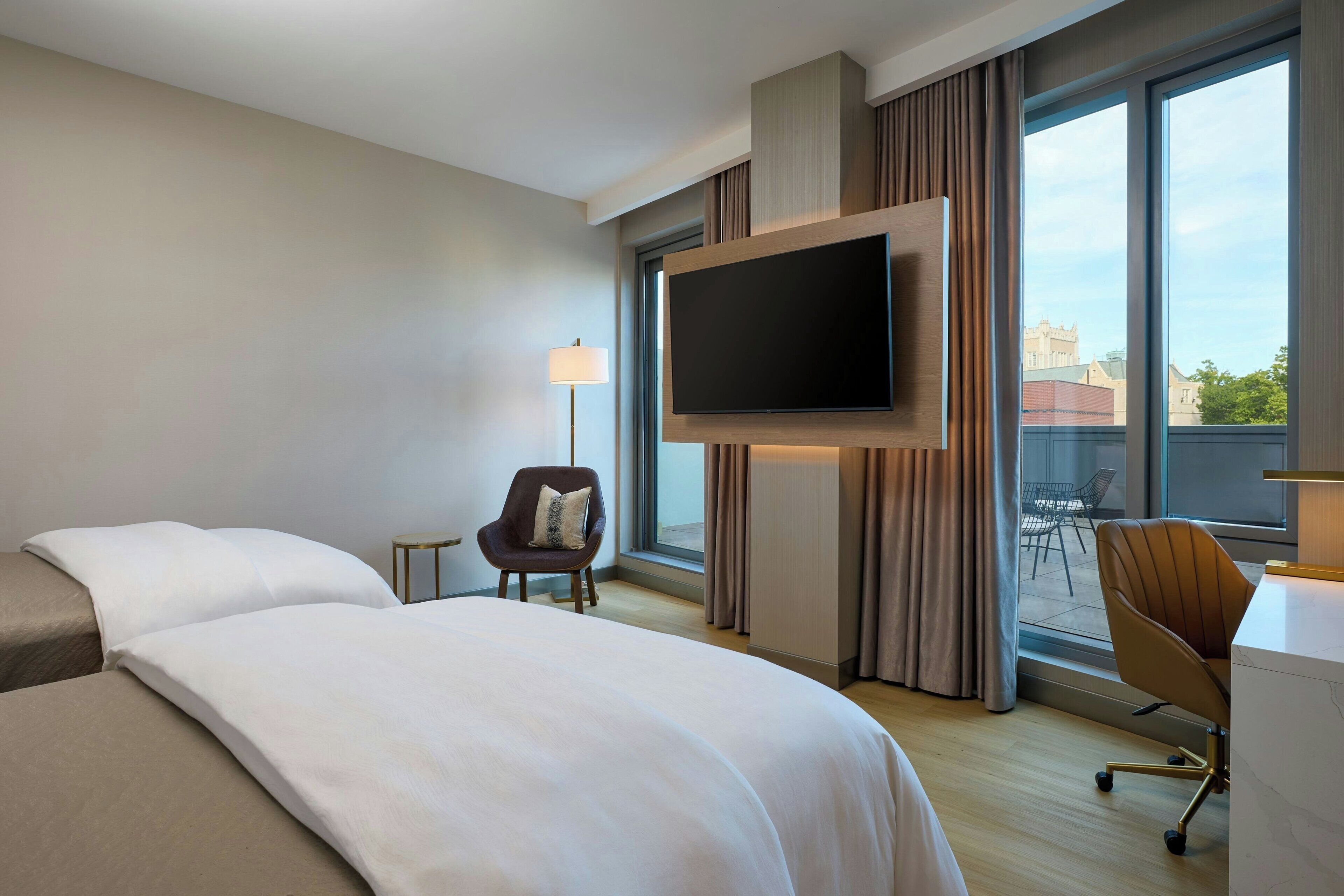 Ein Hotelzimmer mit einem großen Bett, einem wandmontierten Flachbildfernseher, einem Schreibtisch mit Stuhl und Blick auf die Stadt durch große Fenster.