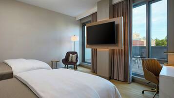 Ein Hotelzimmer mit einem großen Bett, einem wandmontierten Flachbildfernseher, einem Schreibtisch mit Stuhl und Blick auf die Stadt durch große Fenster.