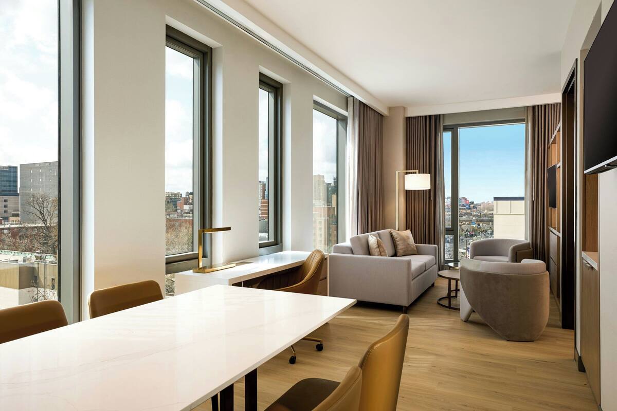 A modern hotel room with a large window, a sofa, a chair, and a dining table.