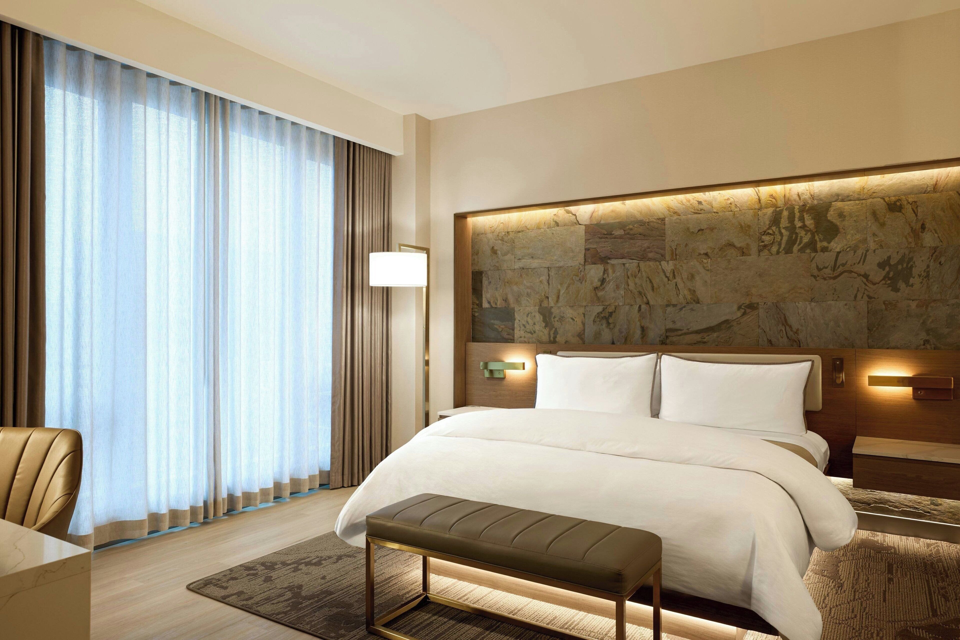 A modern hotel room with a large bed, a bench, a desk, and a view of the city through large windows.