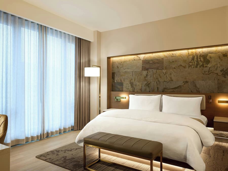 A modern hotel room with a large bed, a bench, a desk, and a view of the city through large windows.