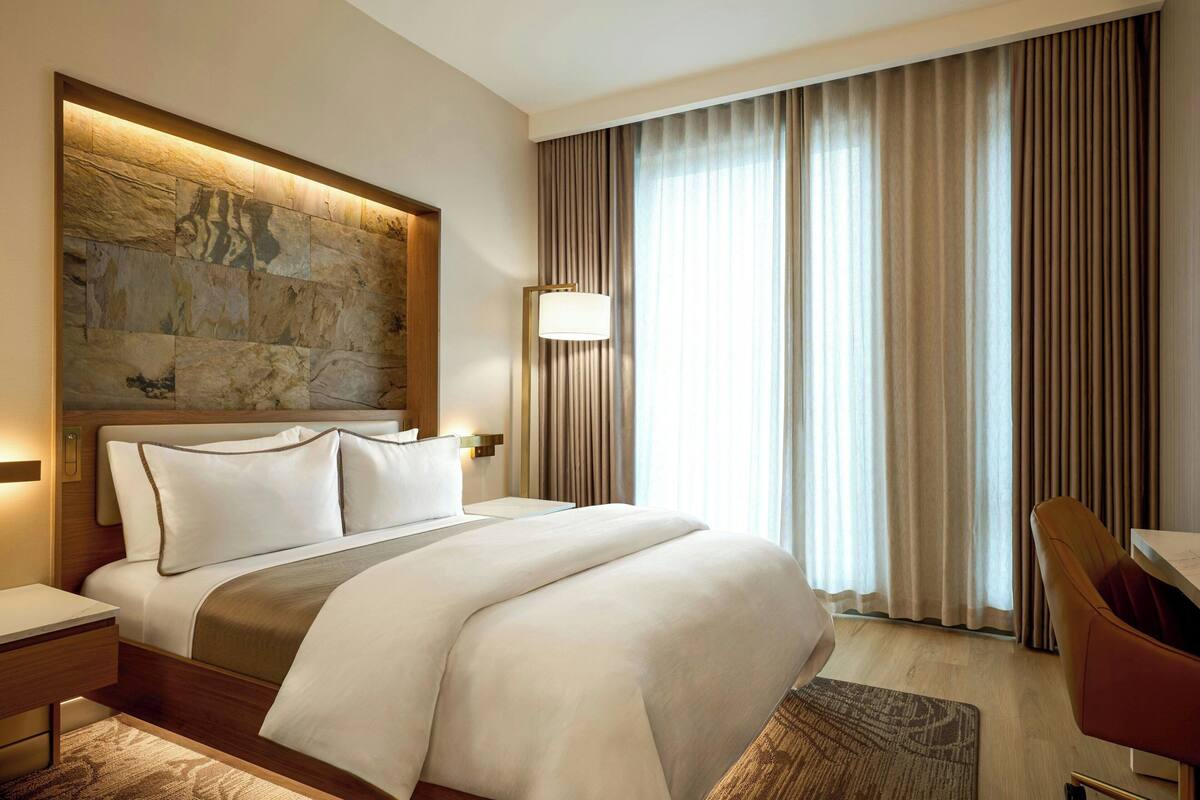 A hotel room with a large bed, a headboard with a stone-like design, a bedside table, a lamp, a chair, and a large window with curtains.