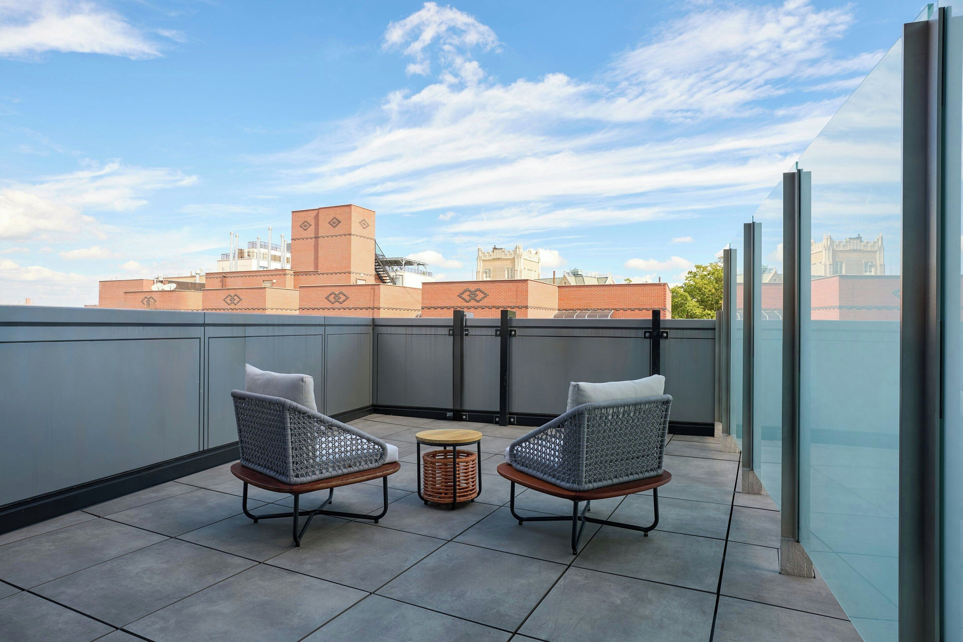 A rooftop terrace with two wicker chairs and a small round table, overlooking a cityscape with buildings and a clear sky.