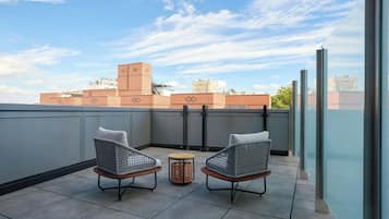 A rooftop terrace with two wicker chairs and a small round table, overlooking a cityscape with buildings and a clear sky.