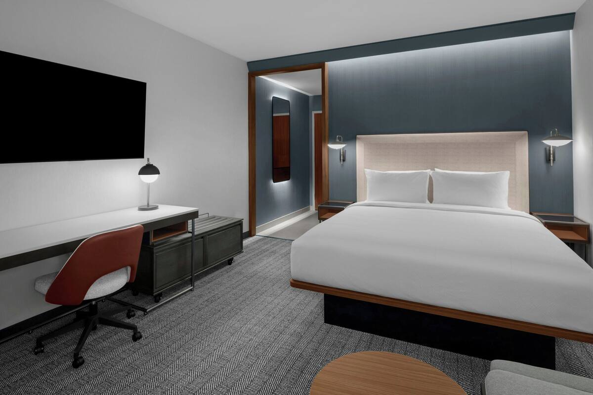 A modern hotel room with a large bed, a desk with a chair, a desk lamp, and a small round table.