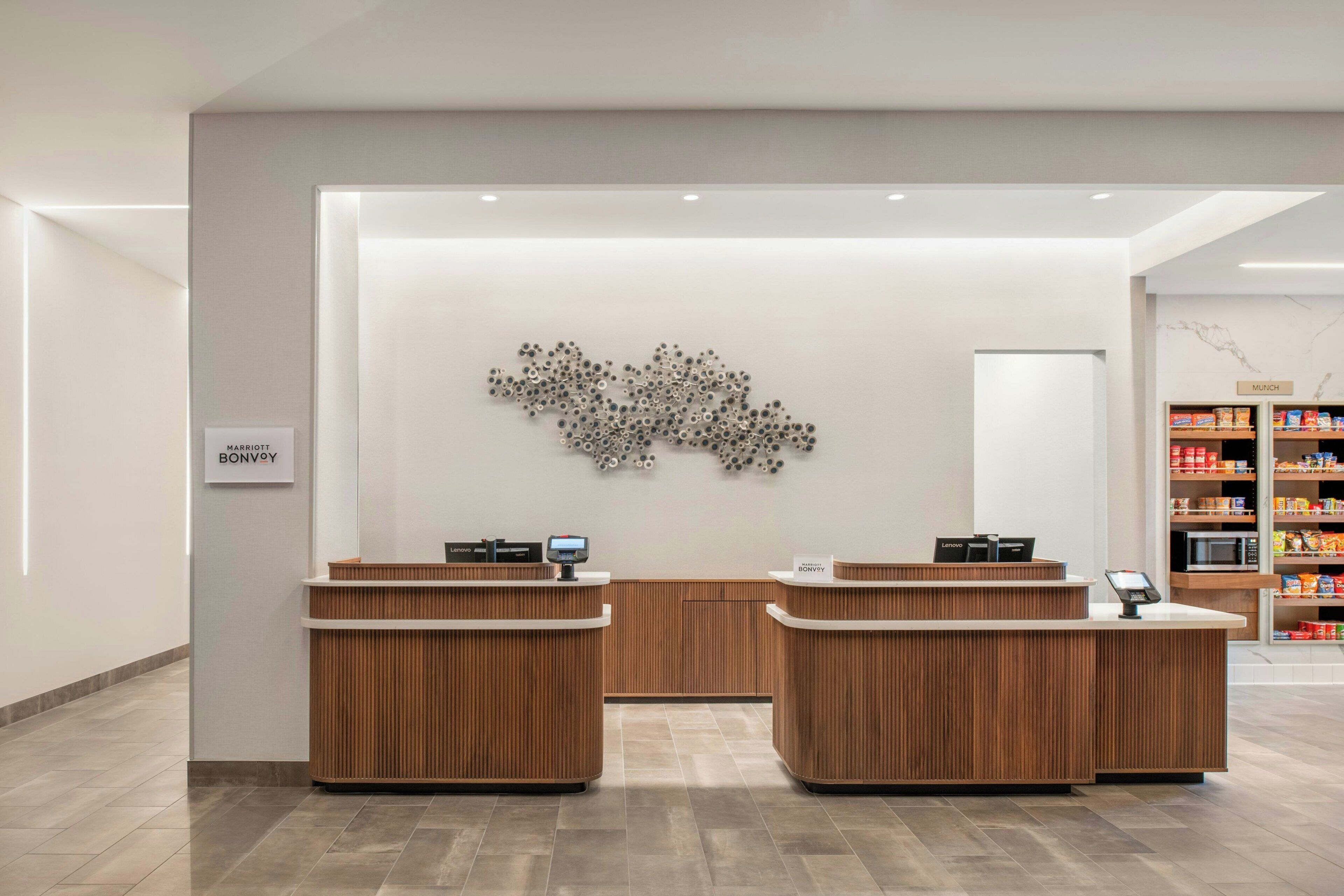A hotel reception area with two wooden reception desks, a wall-mounted artwork, and a self-service kiosk.