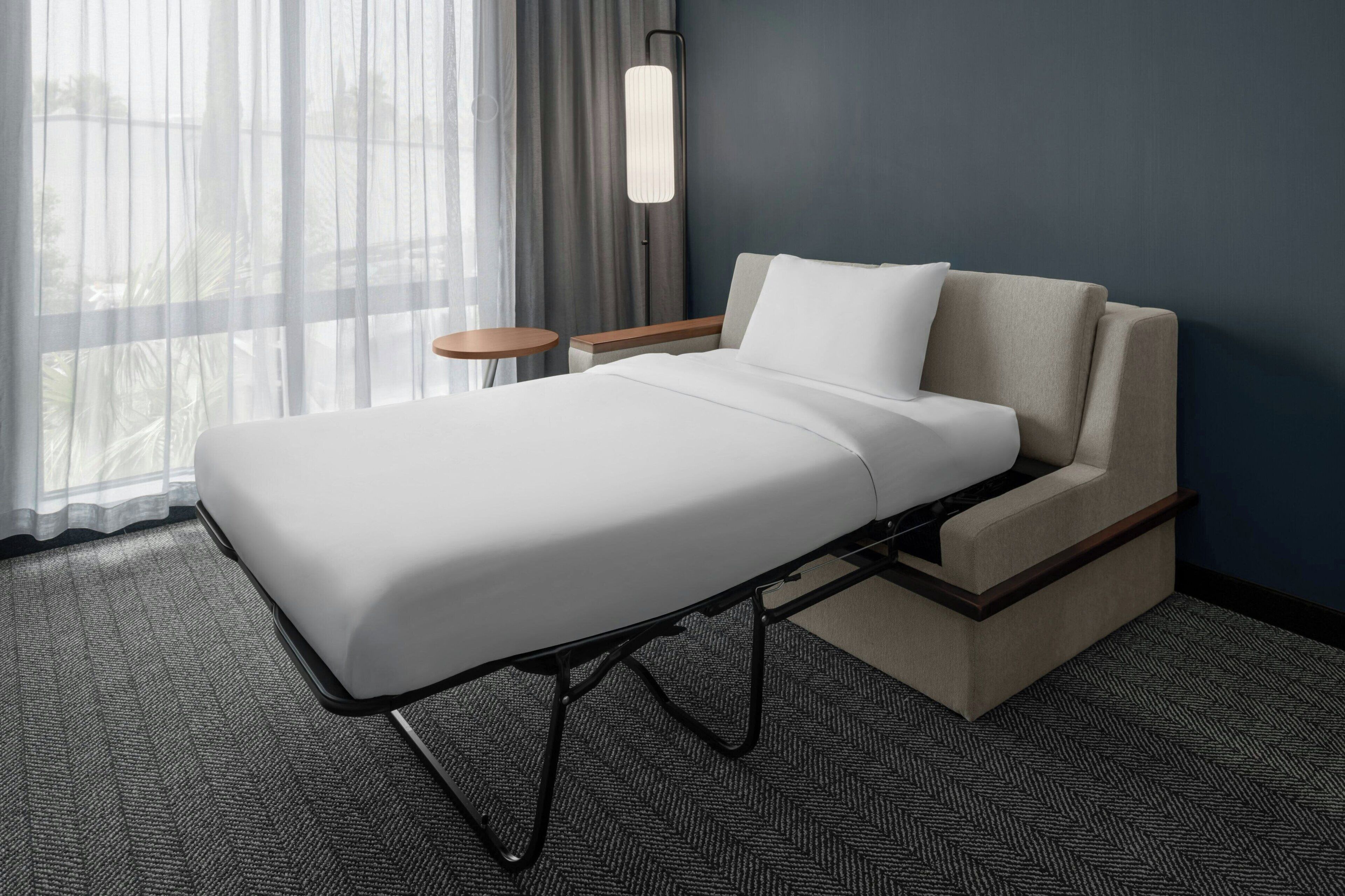 Een moderne hotelkamer met een enkel bed, nachtkastje, lamp en uitzicht door dunne gordijnen.