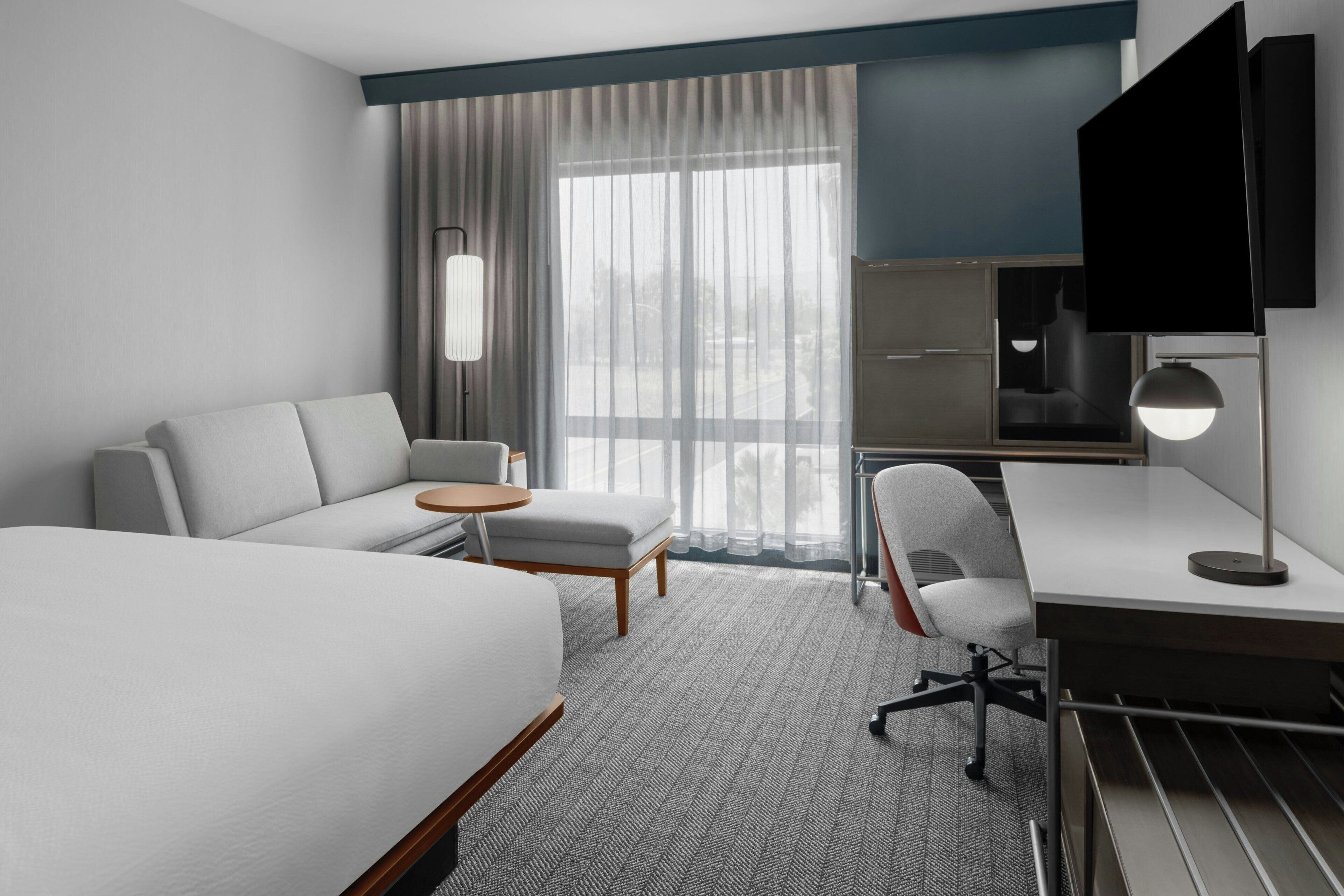 Ein modernes Hotelzimmer mit Bett, Schreibtisch, Stuhl, kleinem Tisch, Fernseher und einem großen Fenster mit Vorhängen.