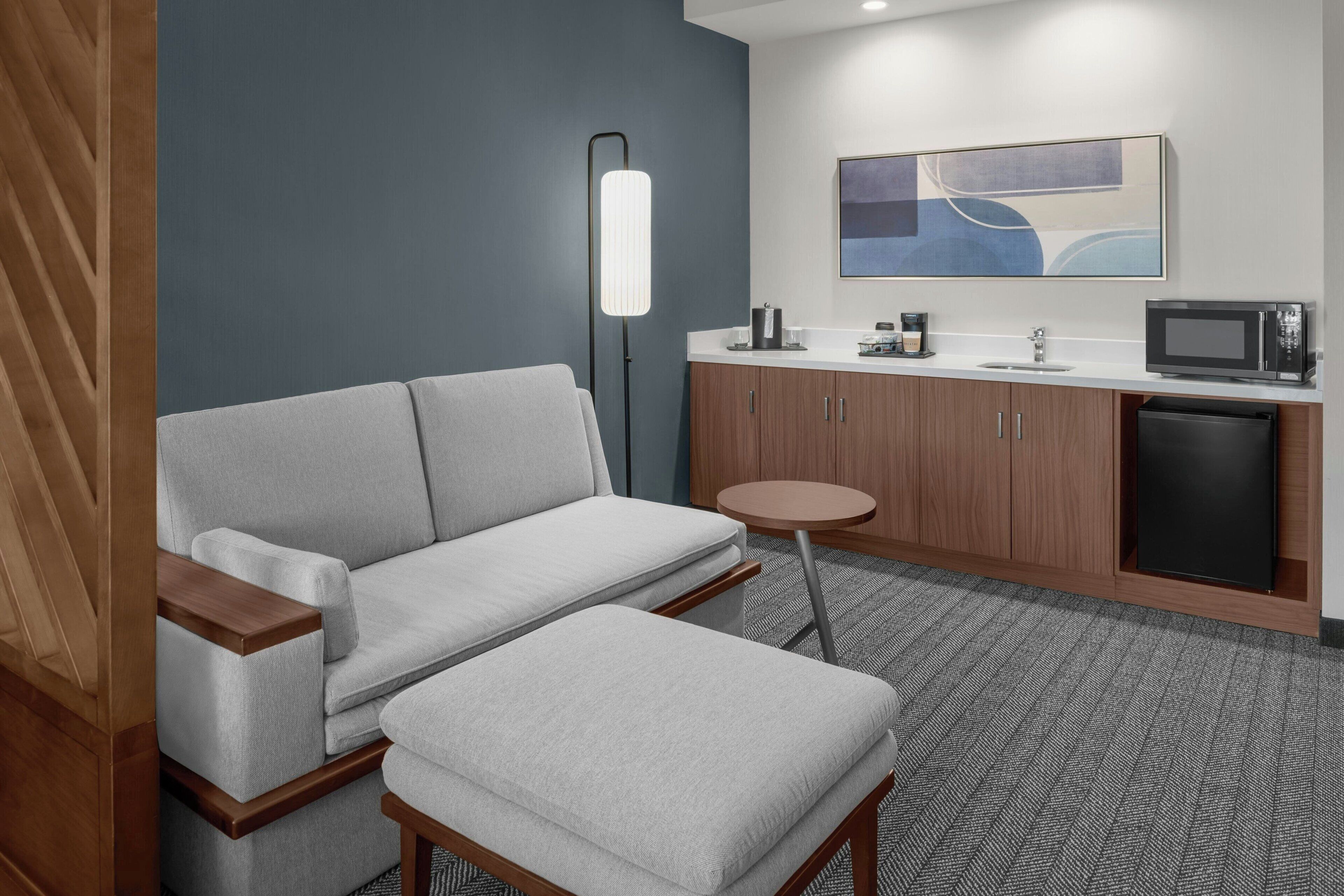 Een moderne hotelkamer met een grijze bank, een klein rond tafeltje, een magnetron en een wastafel.
