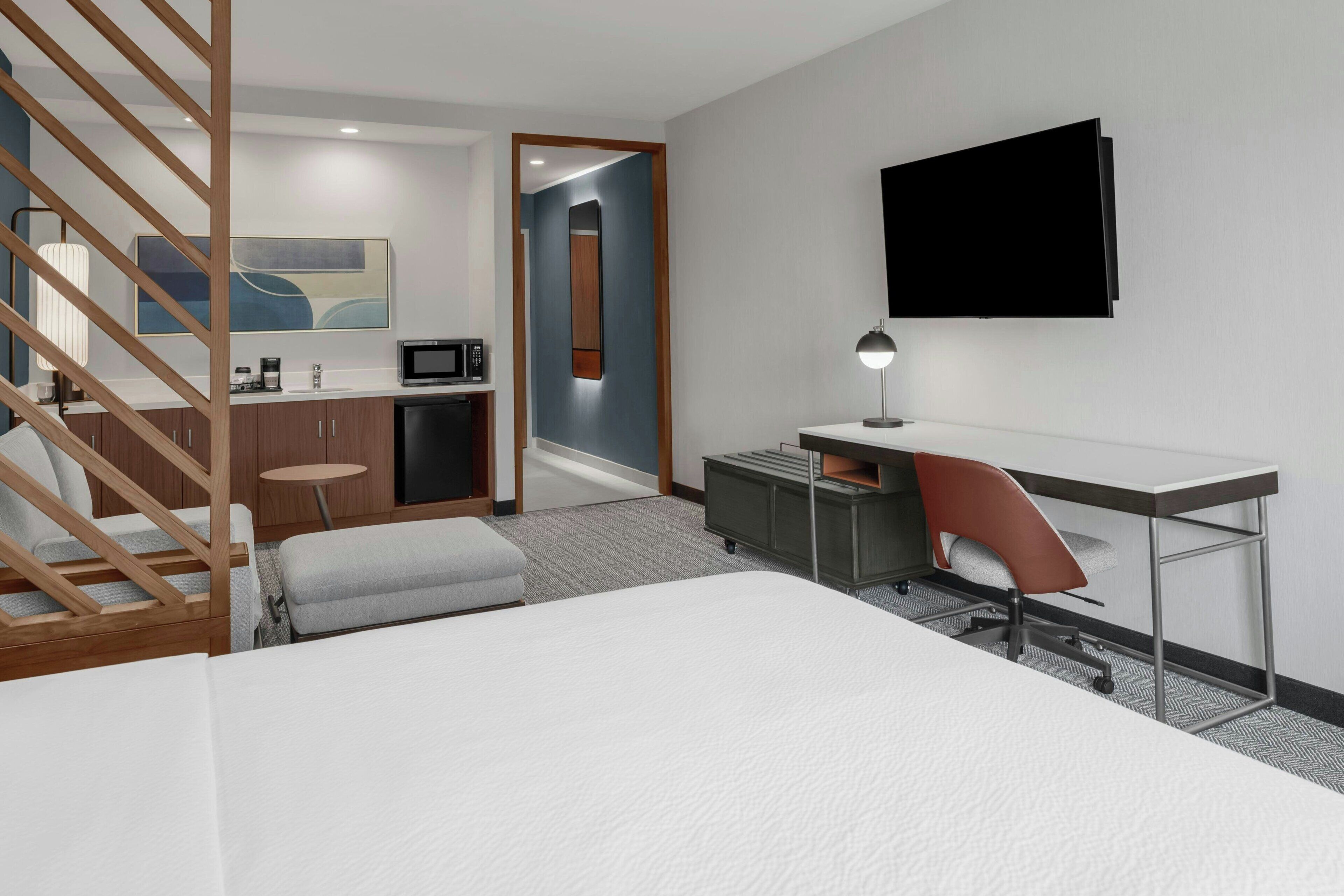 A modern hotel room with a bed, a desk, a chair, a TV, a kitchenette, and a staircase.