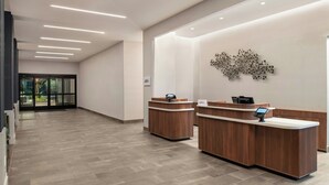 A reception area with two wooden reception desks, a modern light fixture, and a wall-mounted abstract art piece. - Courtyard By Marriott San Bernardino Loma Linda (Loma Linda)