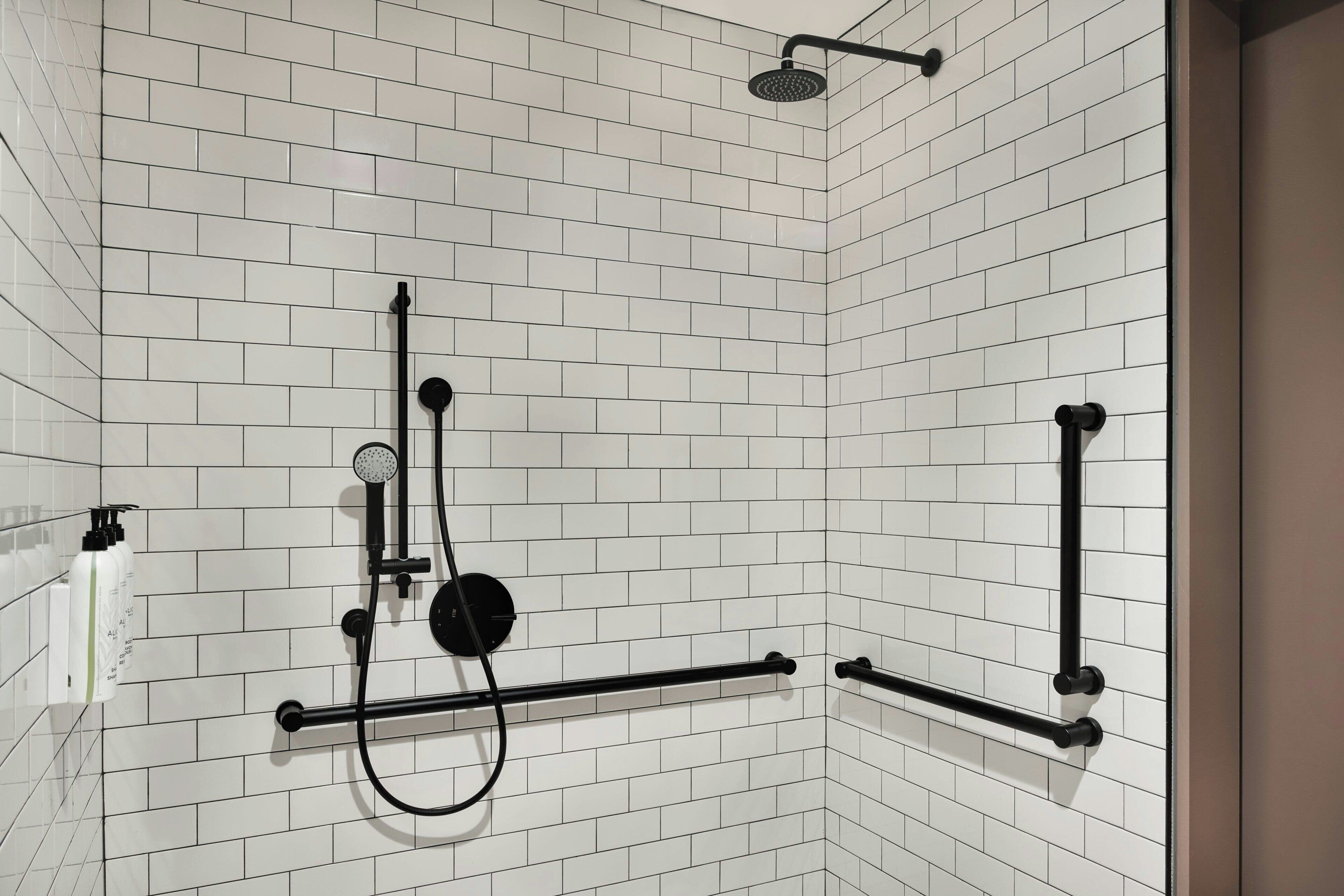 A bathroom with white tiled walls, a shower with a black hose, and a black towel rack.