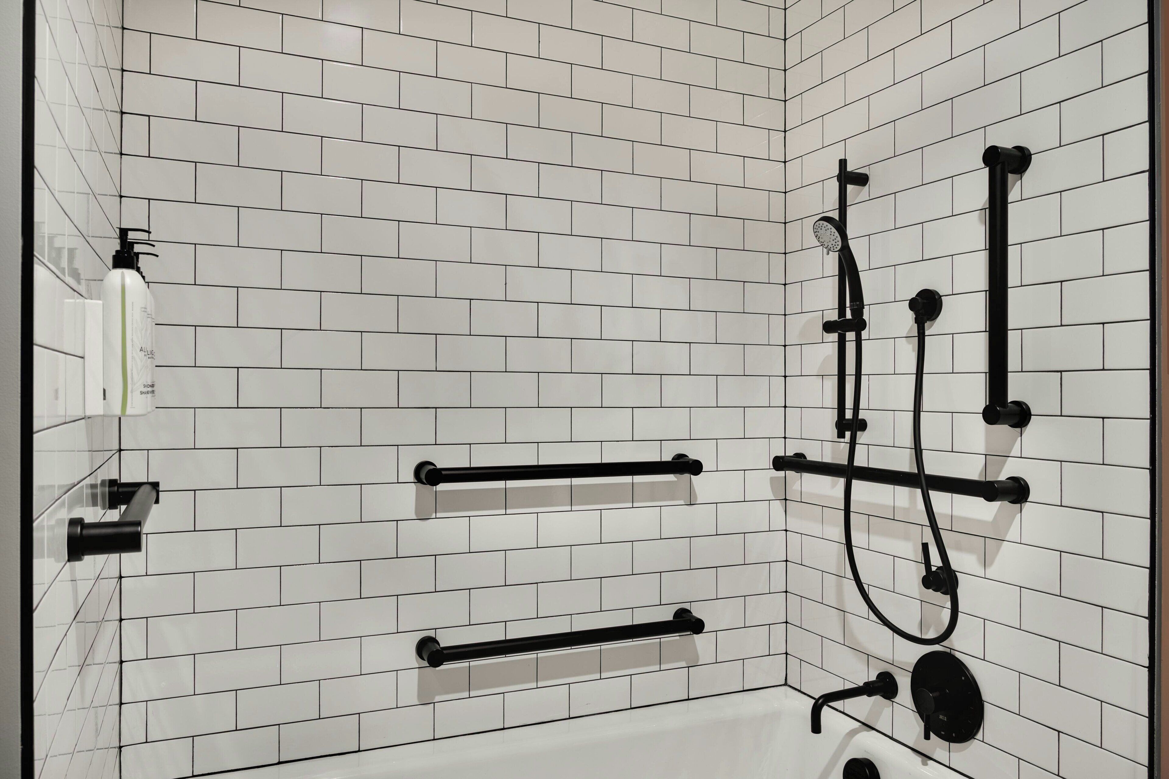 Une salle de bain avec des murs carrelés de carreaux blancs, un porte-serviettes noir, une douchette et un flacon de lotion.