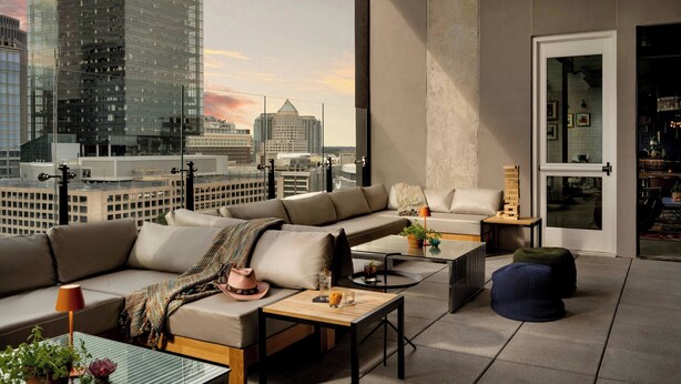 A modern living room with a large window overlooking a cityscape, featuring a sectional sofa, a coffee table, and a small side table with a lamp.