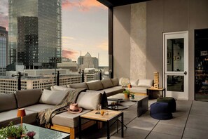A modern living room with a large window overlooking a cityscape, featuring a sectional sofa, a coffee table, and a small side table with a lamp. - Moxy Charlotte Downtown (Charlotte)