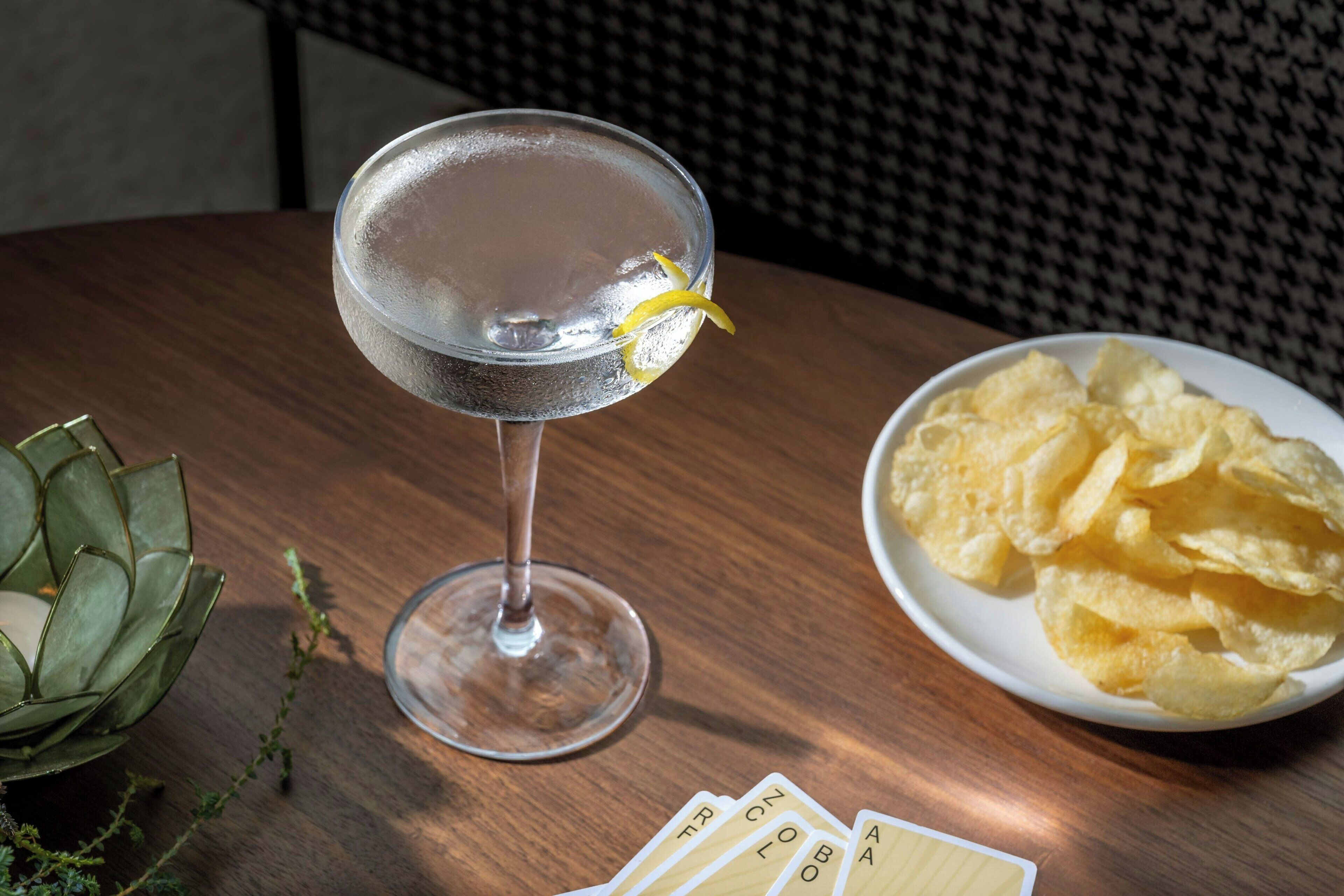 A martini glass with a lemon twist on a wooden table, accompanied by a bowl of chips and playing cards.