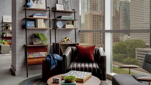 A modern living room with a city view, featuring a leather sofa, a bookshelf with books and decorative items, and a large window.