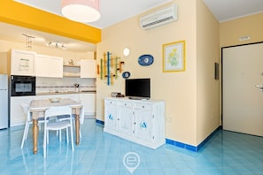 A compact kitchen with white cabinets, a dining table, chairs, a TV on a cabinet, and a wall-mounted air conditioner. - Pine & Sea Hideaway - Torre Grande (Torre Grande)