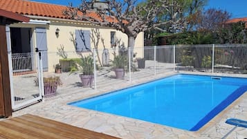 Une piscine avec une terrasse en bois, entourée d’une clôture, et un bâtiment avec un toit en tuiles en arrière-plan.