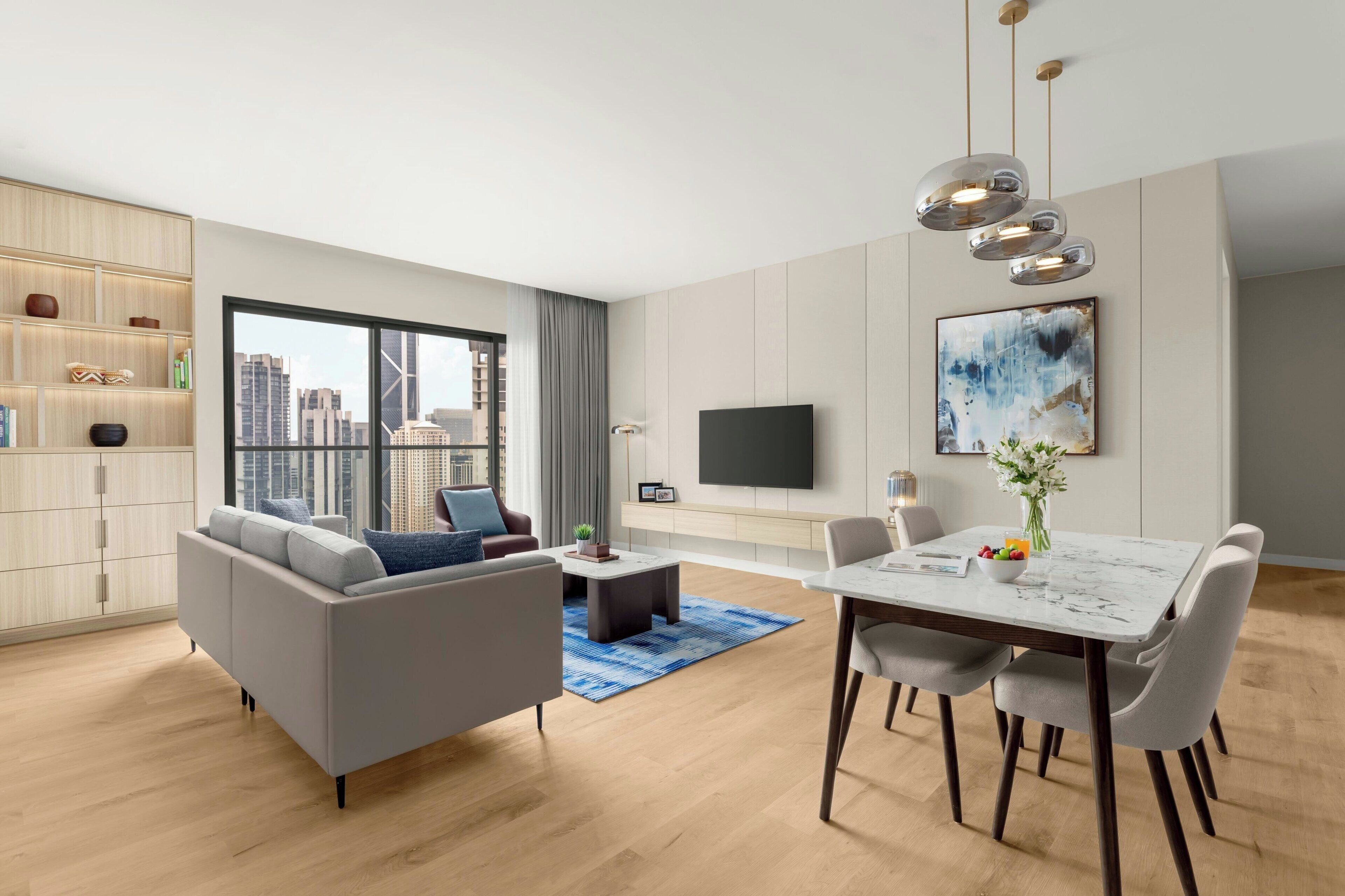 A modern living room with a sofa, coffee table, dining table, and chairs. There is a TV mounted on the wall and a bookshelf with various items.