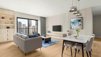 A modern living room with a sofa, coffee table, dining table, and chairs. There is a TV mounted on the wall and a bookshelf with various items.