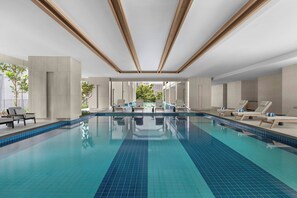 Une piscine intérieure moderne, avec des carreaux bleu clair, des chaises longues et une vue sur la verdure environnante.