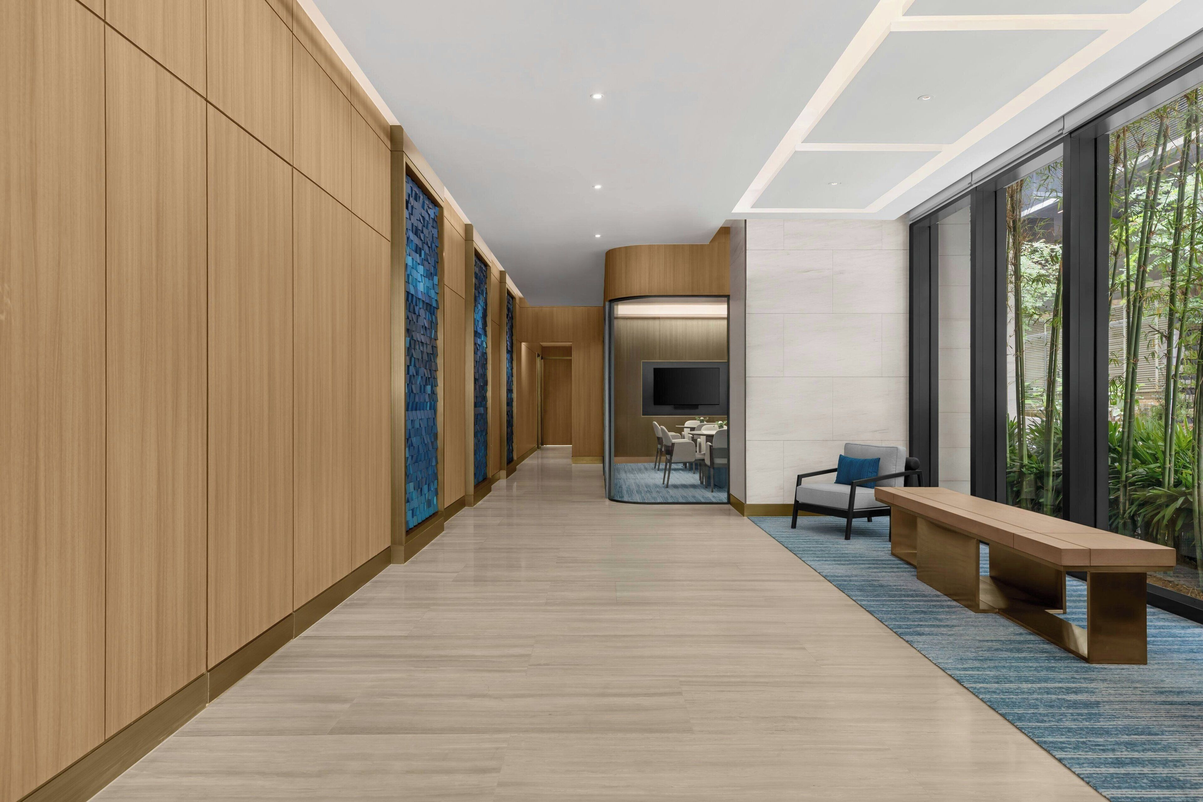 Un couloir moderne avec un lambris en bois, un mur orné d’un motif bleu, un coin salon avec un banc et une grande fenêtre donnant sur un paysage verdoyant.